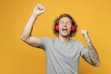 Young expressive cool caucasian man 20s he wearing grey t-shirt wireless headphones dance have fun raise up hands isolated on plain yellow color backround studio portrait. People lifestyle concept