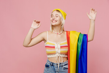 Young fun blond lesbian woman she wear colorful knitted top striped flag on shoulder dance raise up hands close eyes hat isolated on plain pastel light pink background. People lgbtq lifestyle concept.