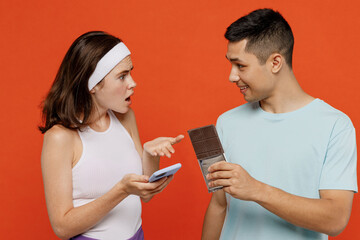 Young fitness trainer instructor sporty two man woman in headband tshirt hold mobile cell phone chocolate count calories spend weekend in gym isolated on plain orange background Workout sport concept
