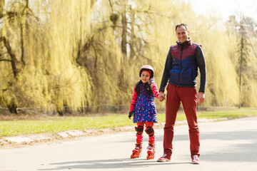 A father teaching his daughter roller skating in a park on summer day. Happy week-end. Father's day