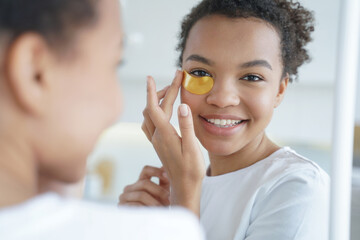 Smiling young mixed race teen girl apply gold under eye patches. Skincare treatment, beauty routine - Powered by Adobe