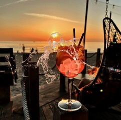  glass of juice with ice water splash and sun beam flares on wooden table top sunset at sea in beach restaurant view in pink sky and sea nature landscape