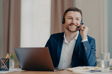 Photo of happy male manager wears headset works on laptop computer consults clients looks with glad...
