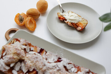 rectangular appetizing homemade apricot pie on a rectangular plate, on a wooden cutting board. Next to it is a round plate with a piece of cake and a spoonful of cream.