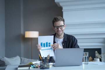 Young smiling german businessman pointing at document with graphics during internet meeting