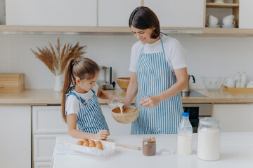 Happy mother and daughter bake together in kitchen, use different ingredients, wear aprons, stand...