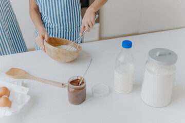Cropped view of kid in apron whisks ingredients in bowl with beater, busy cooking and helping mother to prepare cake. Dough, milk, eggs, wooden spatula and chocolate on table