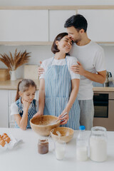 Obraz na płótnie Canvas Happy lovely family in home kitchen, father embraces mother with love, little girl looks in bowl, observes how mommy cooks and whisks ingredients, use eggs for making dough. Domestic atmosphere