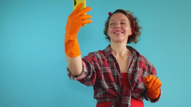 An adult pin-up woman in a plaid shirt and rubber orange gloves takes a selfie on a mobile phone. Portrait of woman cleaning