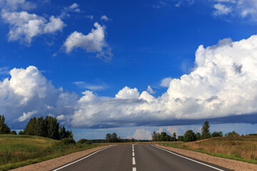 Road up hill with green grass field under white clouds and blue sky.