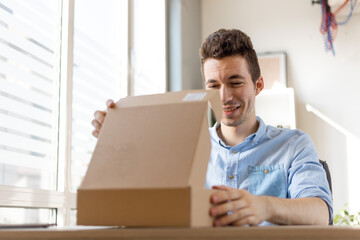 Close up of happy amazed young man consumer holding opening cardboard box sit on sofa at home, order postal shipping courier delivery concept.