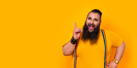 Bearded gay big man with rainbow trouser holders and make-up pointing up with a happy surprise...