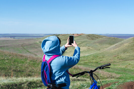 Outdoors view from the back of a young woman in a blue hoodie who is traveling alone on a bicycle in the mountains, stopped to rest to take a photograph of the landscape with her mobile phone camera.