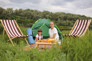 portrait of happy optimistic young woman and little girl sitting on blanket near tent, looking at camera with charming smiles, having picnic, resting near river.