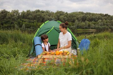 Image of pleased pretty mother having picnic near river with her daughter, sitting on plaid on the ground near tent, talking and enjoying their camping.