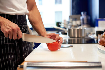 Chef slicing fresh ripe tomatoes on a white chopping board with steel sharp knife. Hands closeup. Professional kitchen. Workshop food concept.