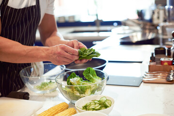 Man chef is preparing green salad of romaine lettuce. Healthy food concept. Cucumber and corn. Copy space. Hands close up. White professional kitchen