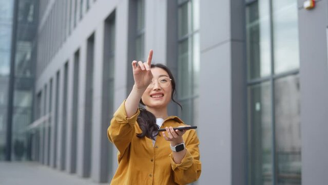 Pleasant chinese woman in casual wear holding modern smartphone and making scroll gesture with finger in air. Dark haired young female touching virtual screen at urban area.