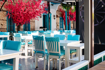 View of summer street cafe with white-blue tables and chairs. Open air restaurant on background of red bougainvillea on the wall with no people. Selective focus.