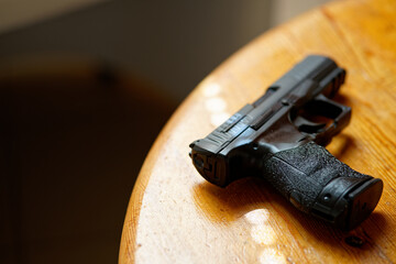black hand gun lays on a wooden table opened blurry doors shadows casted on the floor are in...