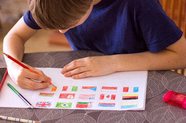 A schoolboy boy draws flags of different countries on a sheet of paper, Ukraine, France, Canada, Germany, Great Britain, Russia