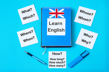 Flat lay blue notepad with text: Learn English and stickers with question words on a blue background. Top view.