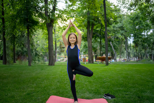 a little girl in the park is doing yoga, she stands on one leg, the other is tucked under her and stretched her arms up