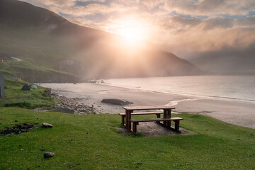 Table and benches for tourist on a grass with stunning view on Keem bay and beach early in the morning at sunrise. Low clouds and fog over ocean and mountain. Ireland. Famous travel area.