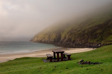 Fototapeta na wymiar Table and benches for tourist on a grass with stunning view on Keem bay and beach early in the morning. Low clouds and fog over ocean and mountain. Ireland. Famous travel area. Irish landscape.