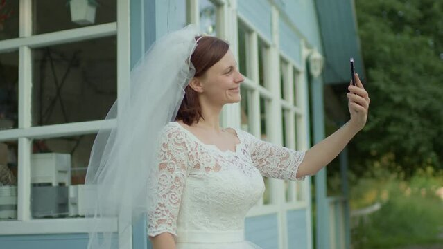 30s bride in white wedding dress using mobile phone standing near rural house. Happy newlywed holding cell phone. Slow motion, handheld camera.