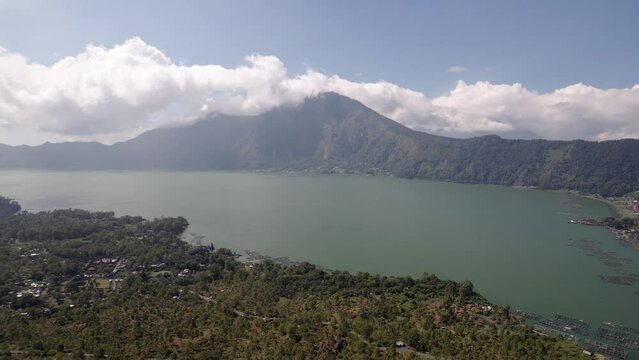 Green Batur lake and Mount Abang in white clouds, aerial shot of Bali island highlands. Village and farmland seen down at lake shore, fish cages floating on water surface