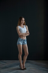 young woman in a white tank top and denim shorts on a dark background, full length portrait