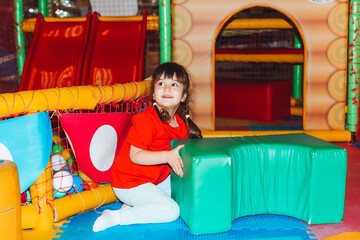 A happy little girl is having fun in an indoor play center. A child plays with colored balls in a pool with a ball on the playground.