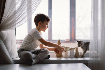 Cute boy playing with kitten sitting on the windowsill near the window. Cozy home with decorative Christmas trees. sweet home. Winter holidays lifestyle. Family lifestyle concept.