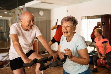 Happy elderly man training on exercise bike and smiling near joyful wife