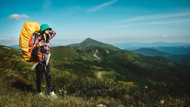 Woman tourist is walking on a hiking trail with a backpack against background of green mountains and sky