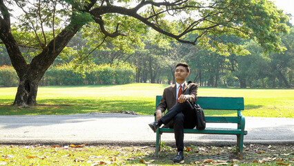 Fototapeta premium Asian young business man sitting on a wooden bench and relaxing in the park