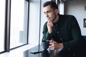 Thoughtful caucasian young businessman with coffee cup looking through window in office