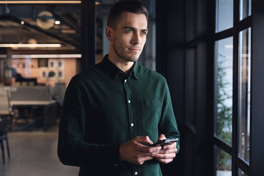 Thoughtful caucasian young businessman with smart phone by window at creative office
