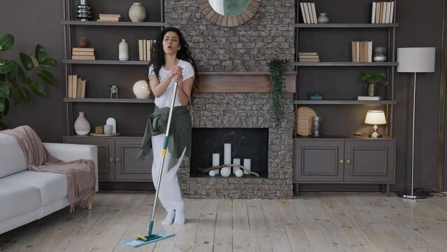 Energetic hispanic woman housewife cleans living room sings song using mop as microphone imagines herself as celebrity on stage attractive girl dancing rhythmic moving to music mopping floor at home