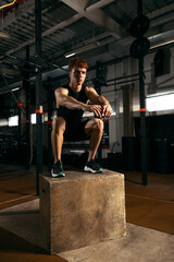 Portrait of sportive, muscular red-haired young man jumping on box, doing exercises isolated over gym background