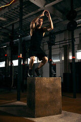 Portrait of sportive, muscular red-haired young man jumping on box, doing exercises isolated over gym background. Strong legs