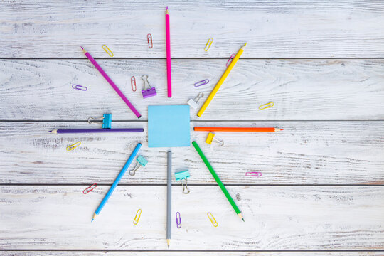 Bright, multicolored pencils for drawing in an album, colored paper clips, a blue notebook for notes and clips are folded neatly in a circle on a light, gray and wooden background.
