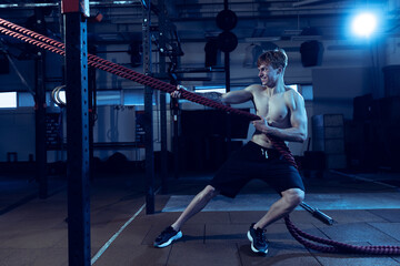 Portrait of muscular red-haired man training, doing full body exercises, pulling ropes isolated over gym background.