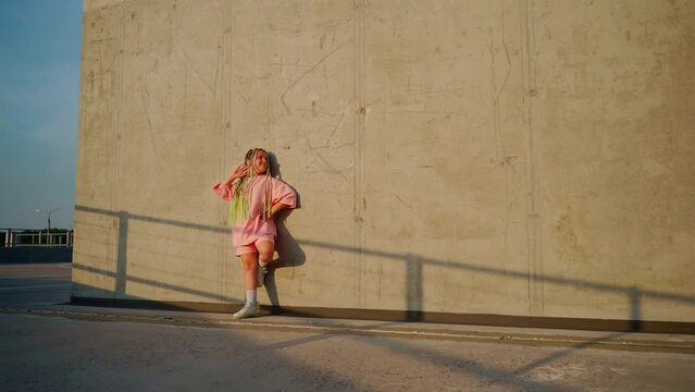 Beautiful Girl Staying At Wall Looking At Camera Outdoors. Caucasian Female Child Portrait Stay Outside. Sun Light At Face. Green Afro Braids Hairstyle And Pink Casual Clothes. Slow Motion Footage