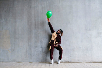 Man with hand raised holding green balloon leaning on concrete wall