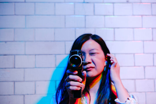 Young woman looking through video camera filming in front of wall