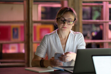 Female employee using mobile device, young woman freelancer distracted by smartphone, sitting at...