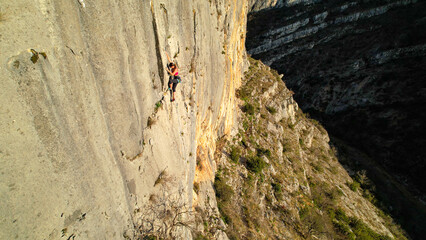 AERIAL: Young female climber lead climbing up the sunlit canyon limestone wall