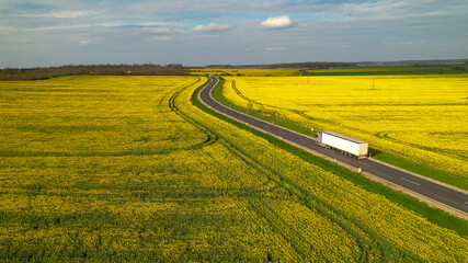 AERIAL: Cargo truck on a delivery mission moving across flowering farm landscape - obrazy, fototapety, plakaty